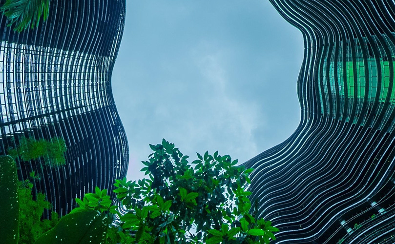 Two modern, curvaceous high-rise buildings with reflective glass facades nestled among green foliage against a cloudy sky