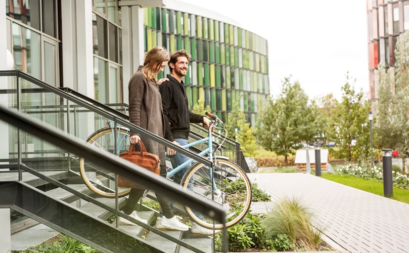 A man with a bicycle and a woman with a brown bag walking down the stairs surrounded by green trees and modern buildings