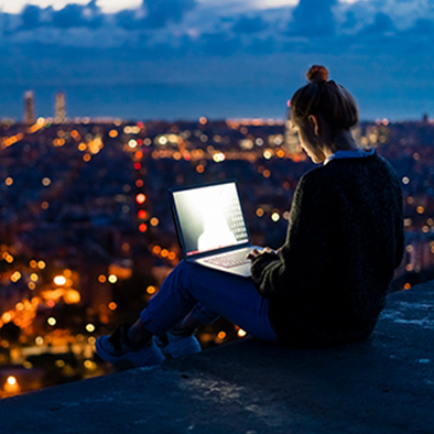 A woman sitting on a ledge with a laptop, with a night-time cityscape in the background